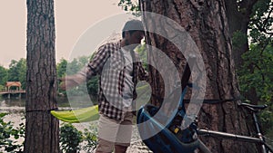 Male cyclist attaches hammock strap to tree in clearing in forest by lake on bicycle trip. Traveler on bike sets green