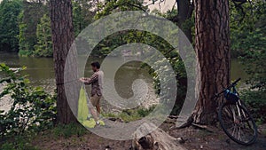 Male cyclist attaches hammock strap to tree in clearing in forest by lake on bicycle trip. Traveler on bike sets green