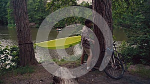 Male cyclist attaches hammock strap to tree in clearing in forest by lake on bicycle trip. Traveler on bike sets green
