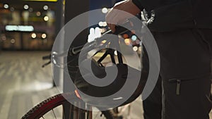 Male cyclist parked his bicycle against pole and hangs his helmet with red light on handlebars of his bike in evening in