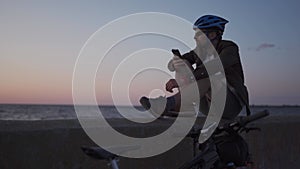 Male cyclist resting on promenade by sea and uses smartphone at sunset. Healthy active lifestyle traveler on bike