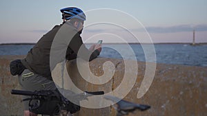 Male cyclist resting on promenade by sea and uses smartphone at sunset. Healthy active lifestyle traveler on bike