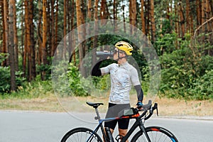 A male cyclist in sportswear stands with a bicycle outside the city and drinks water from a bottle on a background of forest
