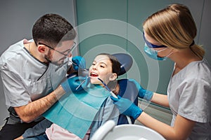 Male dentist check up girl`s teeth with dentist`s tools. Female helper stand beside. Girl sit in dental chair in room