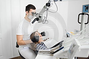 Male dentist with dental tools - microscope, mirror and probe checking up patient teeth at dental clinic office