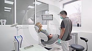 Male dentist preparing young woman for whitening teeth in modern dental office. Doctor adjusts dental chair with female