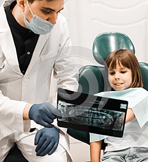 Male dentist showing teeth x-ray to child girl at dental clinic office.