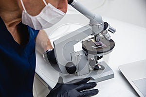 A male doctor in blue lab suit, white hat, mask and black gloves studies a stained histology slide with a microscope