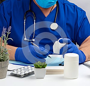 Male doctor in a blue uniform sits at a white table and writes in a paper notebook, wearing sterile gloves on his hands