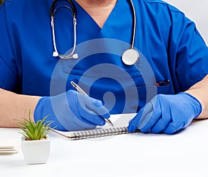 Male doctor in a blue uniform sits at a white table and writes in a paper notebook, wearing sterile gloves on his hands