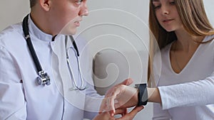 Male doctor explains to patient how to use smart Watch to monitor health and sit on couch in hospital`s office.