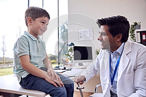 Male Doctor Or GP Wearing White Coat Examining Smiling Boy Testing Reflexes