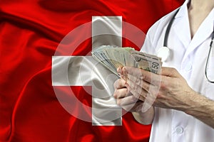 Male doctor holding a fan of dollars banknotes in his hands against the background of the silk national flag of Switzerland,