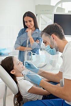 Male doctor with instruments in the hands looking girls teeth sitting in the dental chair while mother standing near her for