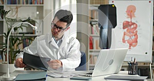 Male doctor looking at x ray photo and shaking head while sitting at table with opened laptop. Man in glasses and white