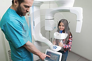 A male doctor makes an x-ray of the girl`s jaw, which sits in a special X-ray machine. The girl is smiling.