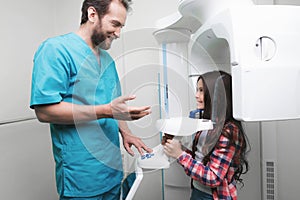 A male doctor makes an x-ray of the girl`s jaw, which sits in a special X-ray machine. The girl is smiling.