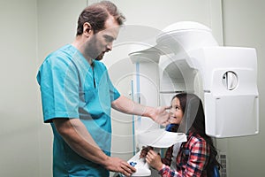 A male doctor makes an x-ray of the girl`s jaw, which sits in a special X-ray machine. The girl is smiling.