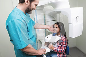 A male doctor makes an x-ray of the girl`s jaw, which sits in a special X-ray machine. The girl is smiling.