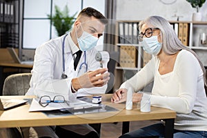 Male doctor in mask measuring temperature of senior woman