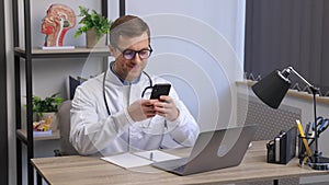 Male doctor medical worker in modern clinic wearing white coat uniform using cell mobile smartphone apps, sitting at laptop