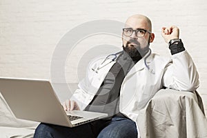 A male doctor sits on a sofa and works in front of a laptop. Remote work, online consultations during the epidemic