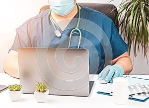 Male doctor sitting at a white table in blue uniform and latex gloves, specialist works on a laptop