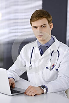 Male doctor sitting and working with laptop in clinic at his working place. Blond physician at work. Perfect medical