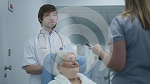 Male doctor stands in clinic corridor with elderly woman, talks to colleague