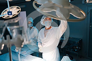 Male doctor in surgical uniforms and masks adjusting light before operation in dark operating theatre. Surgery, hospital