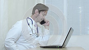 Male doctor talking on the phone, physician working in an office at his laptop.