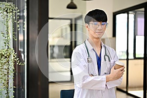 A male doctor in a uniform with a stethoscope stands in the hospital corridor with a coffee cup