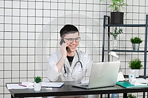 Male doctor using telephone while working on computer at table in clinic