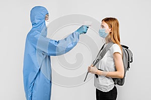 Male doctor wearing protective suit, medical mask with glasses and gloves, uses infrared forehead thermometer to check body