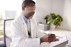 Male Doctor Wearing White Coat In Office Sitting At Desk Working On Laptop