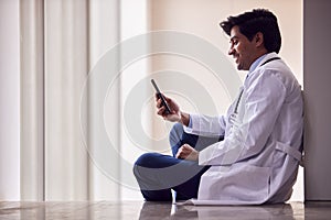 Male Doctor Wearing White Coat Sitting On Floor In Hospital Corridor Text Messaging On Mobile Phone