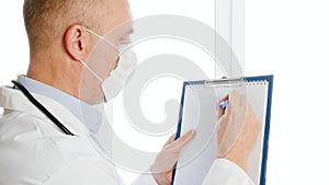 Male doctor in white coat and protective mask writing coronavirus on clip board in hospital. Virologist in medical gown