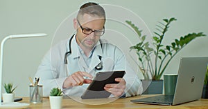 Male doctor in white coat using modern TabletPC device with touch screen. Doctor using DigitalTablet texting to patient