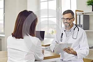 Male doctor writing prescription or making notes in patient's card during medical consultation.