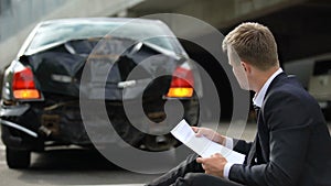 Male driver sitting on road, looking through insurance contract after car crash