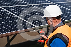 Male electrical engineer in a hard hat with electric wrench maintains solar panels at power plant.