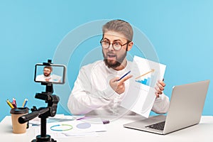 Male employee analyzing graph of clients growth, recording video on smartphone camera or streaming online, conducting conference,