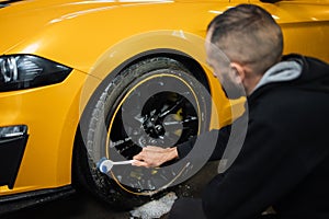 Male employee worker cleaning the wheel tire of modern yellow sport car by scrubbing brush