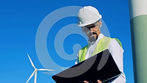 Male energetics worker is standing with his laptop near a wind turbine. Clean, eco friendly energy concept.