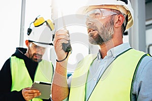 Male engeener wearing protect glasses and helment using walkie-talkie talking in production site. Team of structural engineers