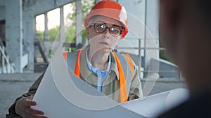 Male engineer talking with a woman in hard hat and safety vest holding blueprint of building, one of them using two way