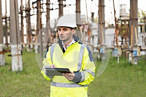 Male engineer in uniform and helmet standing outdoor and tapping on tablet. Man standing near high voltage substation