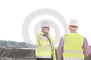 Male engineer using mobile phone while standing with colleague at construction site against clear sky