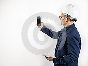 A male engineer in a white hard helmet makes a video call on a smartphone. Middle-aged man in a blue jacket with a tablet computer