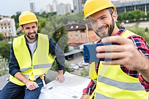male engineers in safety vests and helmets with blueprints taking selfie on smartphone
